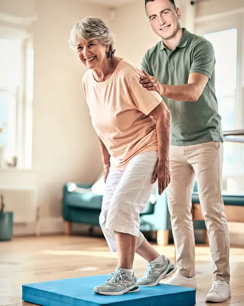 Dans un salon, un physiothérapeute en polo vert sécurise une dame âgée qui s’exerce à l’équilibre sur un coussin de mousse bleu